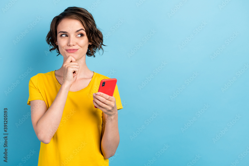 Photo of minded playful lady hold telephone look side empty space finger chin wear yellow t-shirt isolated blue color background Photo of minded playful lady hold telephone look side empty space finger chin wear yellow t-shirt isolated blue color background