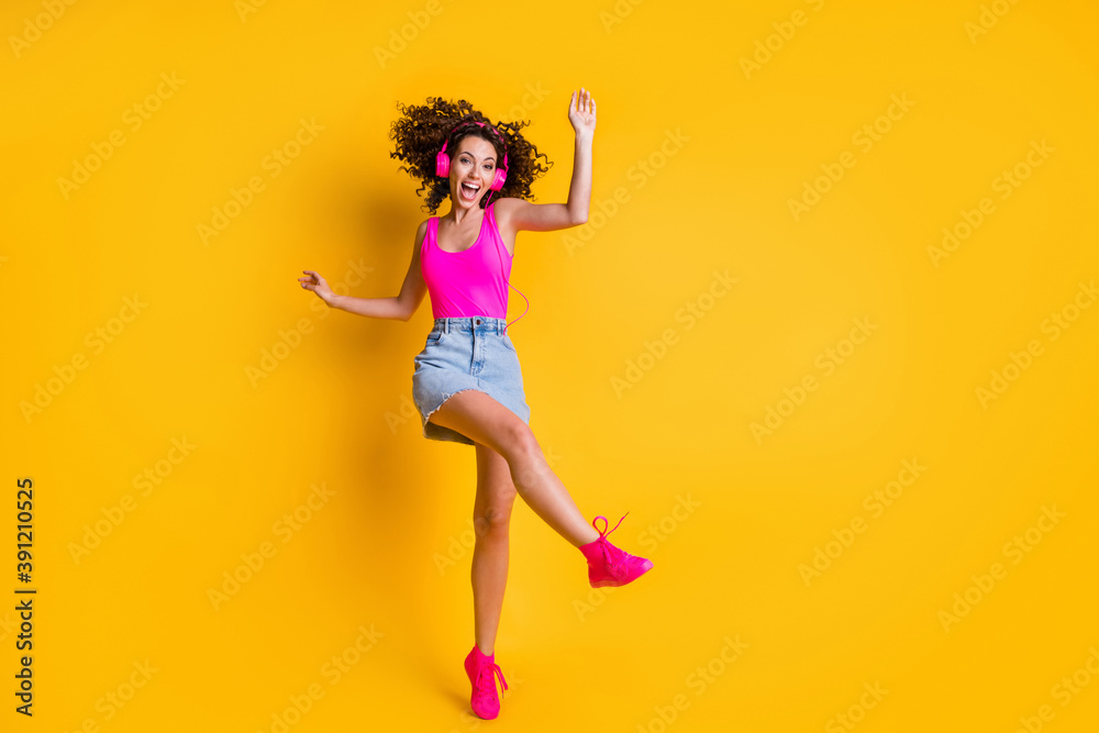 Full body photo of crazy charming curly lady dance carefree raise leg student party delighted listen earphones wear pink tank top denim mini skirt shoes isolated bright yellow color background
