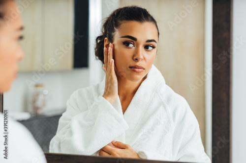 Mixed race woman standing in the bathroom touching her face while looking at a mirror photo