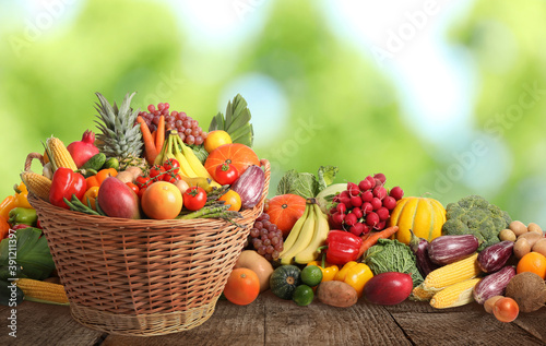 Wicker basket with different fresh organic vegetables and fruits on wooden table against blurred green background
