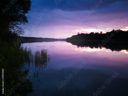 Beautiful summer landscape of a calm river with reflection, gloomy clouds with showers and mountain peaks in the distance during the evening. The setting sun turns the clouds purple