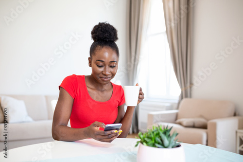 Young woman using a smartphone and having coffee at home. Young african american woman drinking coffee and chatting with friends at social network with her mobile phone