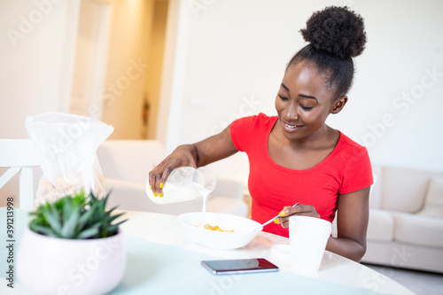 Smiling young African American woman pours corn flakes in plate with milk. The girl has a healthy breakfast on stylish cozy home at the morning while checking her email on laptop.