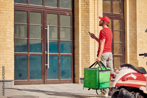 Young male courier in red uniform with thermo bag using his smartphone while standing near customers house on the street