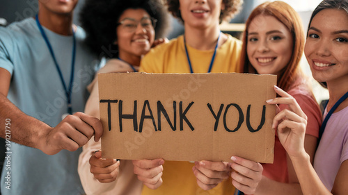 Close up of Thank you inscription, Group of diverse young volunteers smiling at camera, holding card with lettering while standing in charitable organization office © Svitlana