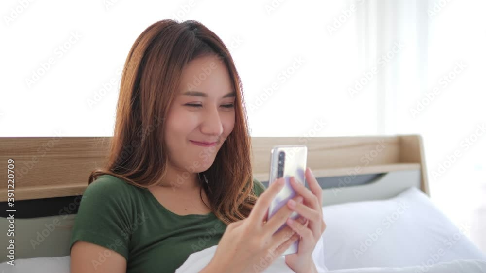 Beautiful Asian Woman using mobile phone enjoying chatting, surfing on the internet on a phone on bed. Happy woman looking at smartphone texting messages, chatting and smile at bedroom. Beautiful Asian Woman using mobile phone enjoying chatting, surfing on the internet on a phone on bed. Happy woman looking at smartphone texting messages, chatting and smile at bedroom.