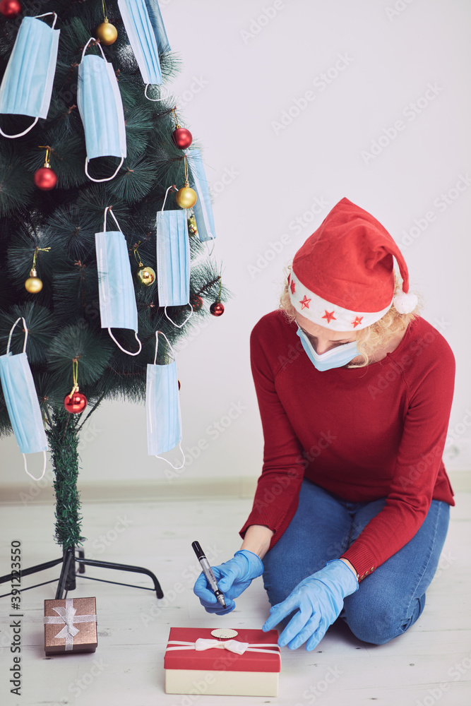 Woman with gift box present, decorating christmas tree with medical masks during virus pandemic 2020 / 2021.