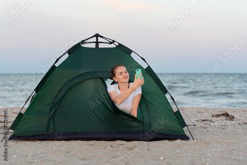 Smiling young woman in tent on seashore. Take photo on smartphone