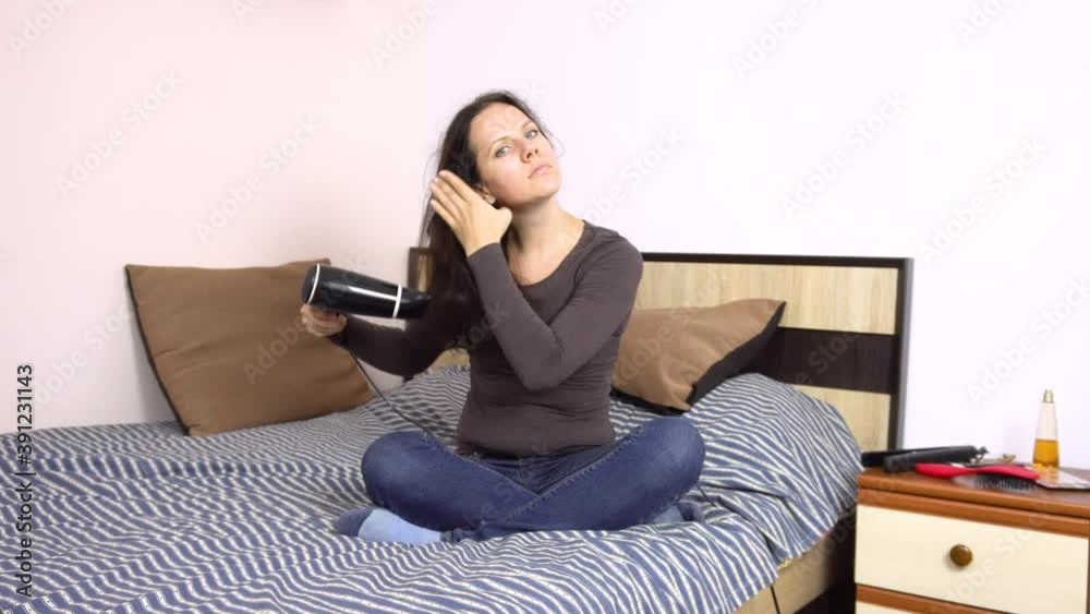 Young beautiful woman dries her hair with a hairdryer on a large bed in her room.Hygiene. Hair styling.4k Young beautiful woman dries her hair with a hairdryer on a large bed in her room.Hygiene. Hair styling.4k