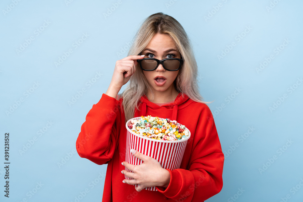 Teenager girl over isolated blue background surprised with 3d glasses and holding a big bucket of popcorns