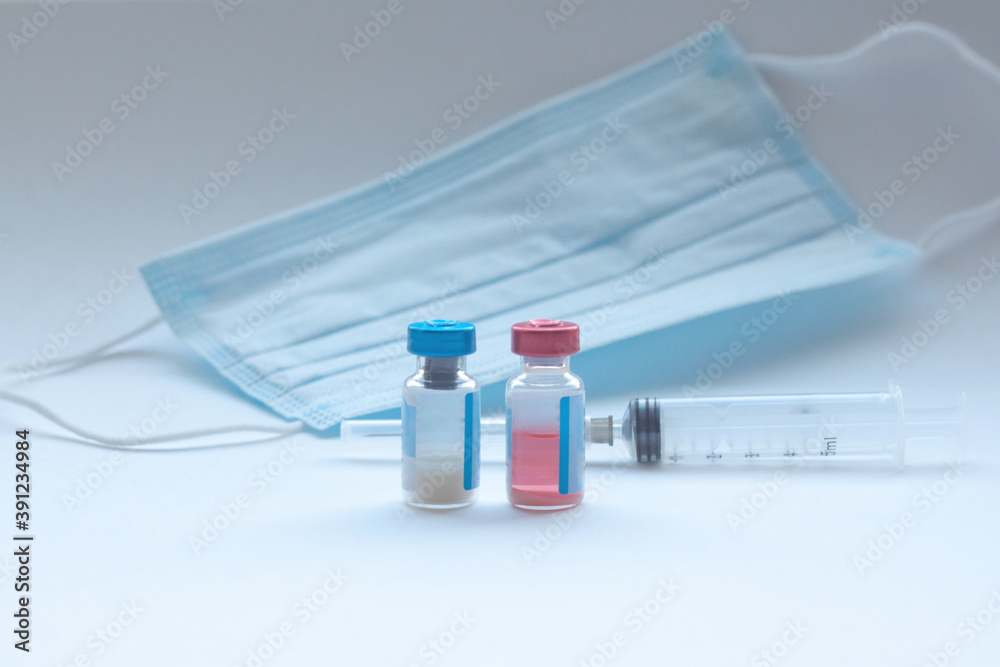 Two vials of vaccine stand on a white background with a medical mask and a syringe in the background. The concept of vaccination, the invention of the vaccine, a new reality, pandemic, kovid-19 Two vials of vaccine stand on a white background with a medical mask and a syringe in the background. The concept of vaccination, the invention of the vaccine, a new reality, pandemic, kovid-19