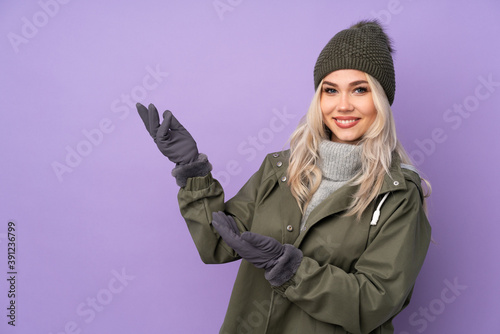 Teenager blonde girl with winter hat over isolated purple background extending hands to the side for inviting to come