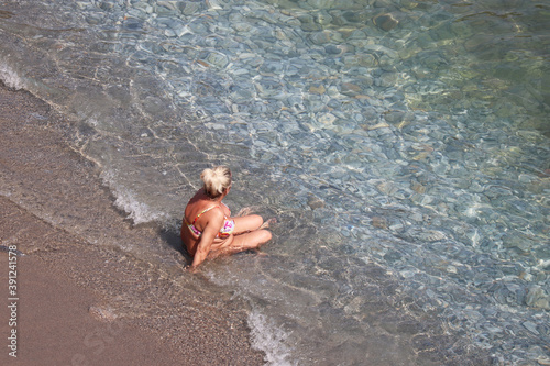 Woman with big breast and fat belly tanning on a beach sitting in sea waves, top view. Vacation, overweight and obesity concept photo