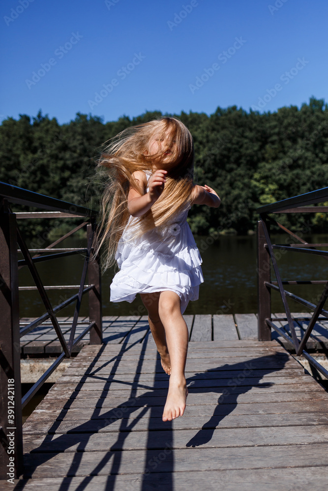 A six year old girl is wearing a white dress. Beautiful long hair. Walk near the river on the background of the forest