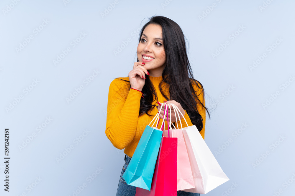 Young Colombian girl over isolated blue background holding shopping bags and thinking Young Colombian girl over isolated blue background holding shopping bags and thinking