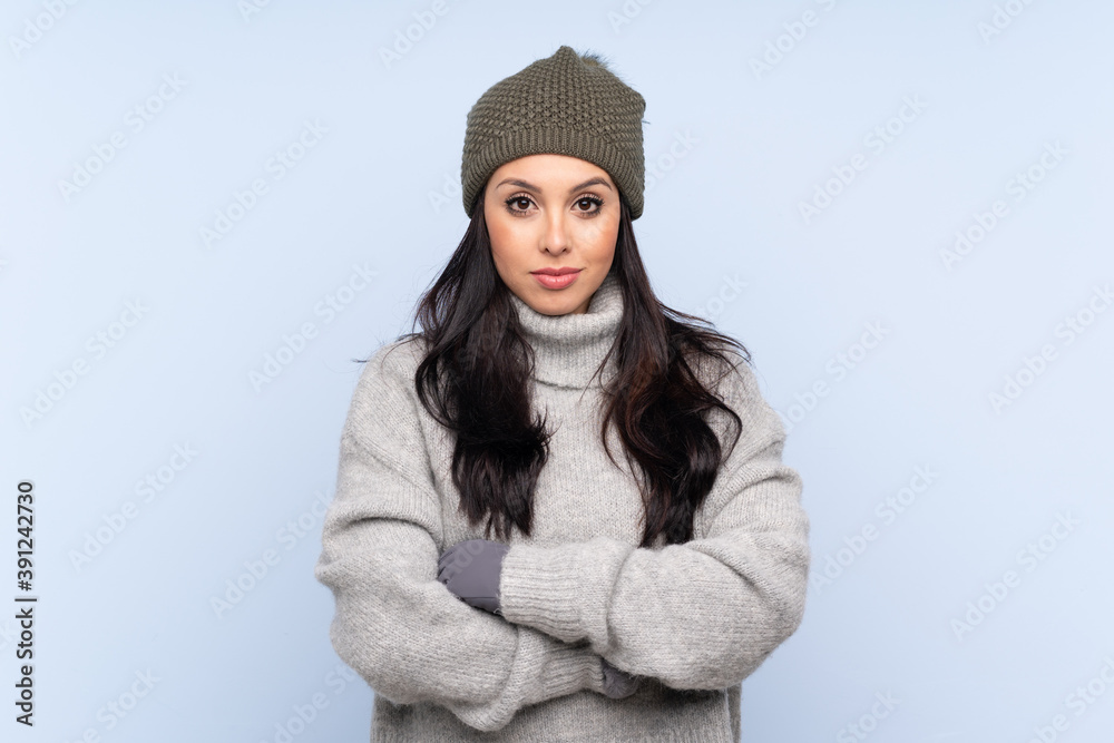 Young Colombian girl with winter hat over isolated blue background keeping arms crossed Young Colombian girl with winter hat over isolated blue background keeping arms crossed