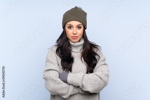 Young Colombian girl with winter hat over isolated blue background keeping arms crossed