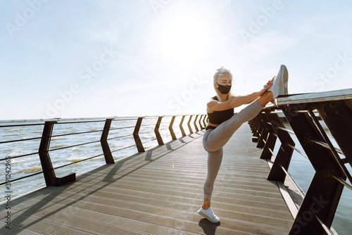 Healthy woman training in protective mask on seaside promenade. Young woman in sportswear does leg stretching before exercise on the beach at sunrise. Morning workout. Covid-19.
