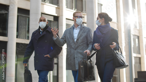 Group of colleagues in formal suit and safety mask walking together past city building outdoors