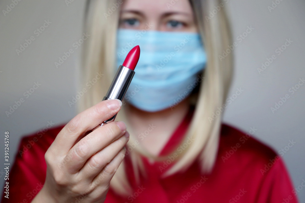 Woman in a protective medical mask with red lipstick in her hand. Makeup and cosmetics during the coronavirus pandemic Woman in a protective medical mask with red lipstick in her hand. Makeup and cosmetics during the coronavirus pandemic