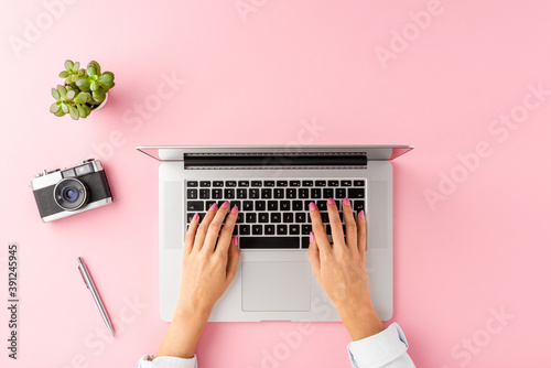 Overhead shot of woman’s hands working on laptop on pink table with accessories. Office desktop. Flat lay