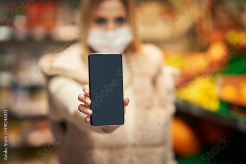 Adult woman in medical mask using smartphone and shopping for groceries