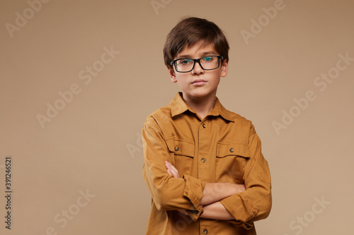Waist up portrait of boy wearing glasses while standing with arms crossed against beige background in studio, copy space