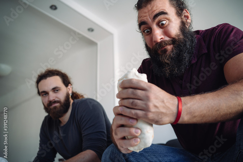 A bearded man looks his durum kebab with a funny expression while sitting on the sofa and his friend looks at him photo