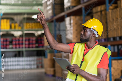 Africa American workers using tablets and checks goods in the automotive parts warehouse distribution center. Male engineers people wear a safety helmet and vest. In background shelves with goods