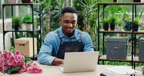 Joyful young man in flower shop sitting at workplace and texting on computer. Cheerful African American male florist worker in floral house tapping and typing on laptop. Floristry concept