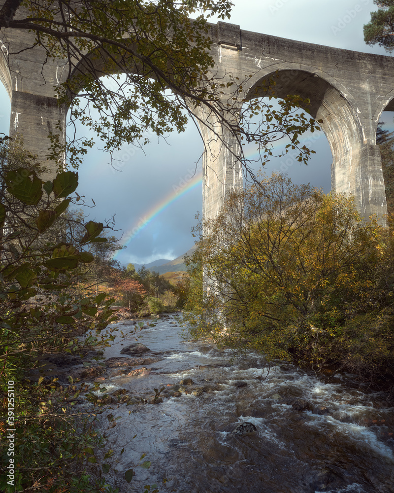 Autumn view of a mountain stream flowing under a big stone viaduct and rainbow after rain. Glenfinnan Viaduct, Scotland, United Kingdom