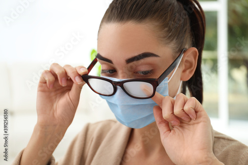 Woman wiping foggy glasses caused by wearing medical mask indoors  closeup