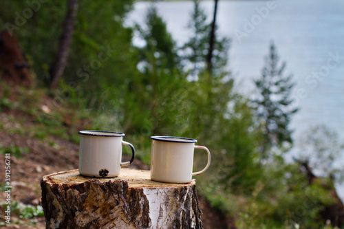Two white metal aluminum enameled mugs with hot tea with steam on a felled wood table on the shore of Lake Baikal in a camp on a natural summer green background.