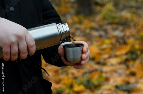 Man pours a hot drink from a thermos into a mug outdoors. Close-up male hands holding a hiking flask against the background of an autumn forest