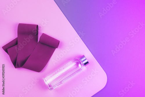 Top view of neon purple and pink colored  maroon expander elastic band, eco reusable glass water bottle on pink  yoga mat with copy space.