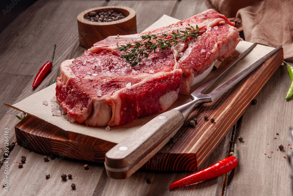 Raw uncooked beef steak. A large piece of meat on the bone lies on a wooden board with spices and fresh herbs.