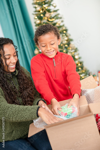 Black mothr helps African American Son Pack Warpped Christmas Presents into Cardbord Shipping Box in Front of Christmas Tree photo