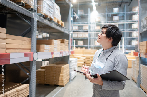 Young Asian man worker doing stocktaking of product in cardboard box on shelves in warehouse by using clipboard and pen. Physical inventory count concept