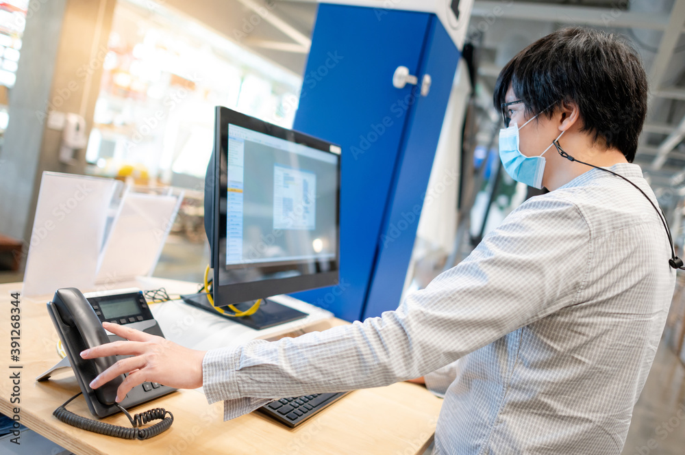 Male Asian customer support operator wearing face mask trying to response customer call by using landline phone on working desk in office. Call center business concept Male Asian customer support operator wearing face mask trying to response customer call by using landline phone on working desk in office. Call center business concept