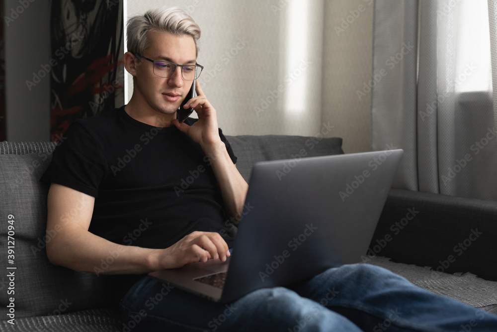 A man with glasses on the couch at home is working on a laptop and talking on the phone. Remote work concept during a pandemic