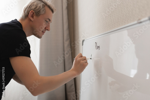 A man with white hair writes "plans" on a white board