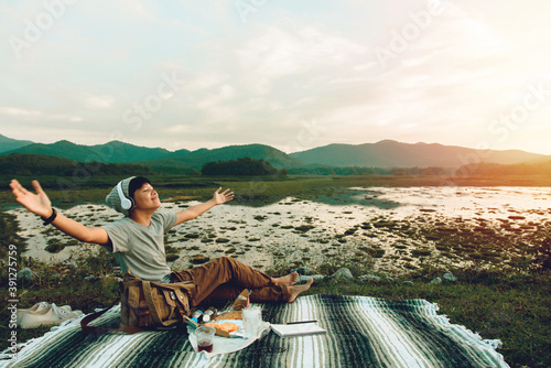 Happy Asian young man in headset listening to music at close range are sitting the wool cloth and see the views with valleys and rivers nature. at MEA TEEP Lamphun in Thailand with sunset background. photo