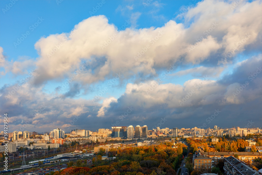 The colorful cityscape of autumn Kyiv, the sun's rays from behind gray clouds catch and brightly illuminate the orange foliage of the city park.