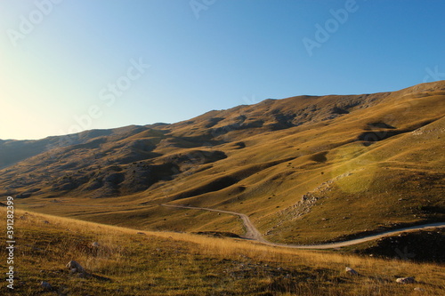 Fototapeta Naklejka Na Ścianę i Meble -  Road through the unevenness of the Bosnian mountain Bjelasnica. The grass in autumn gives a yellow or golden color before sunset.