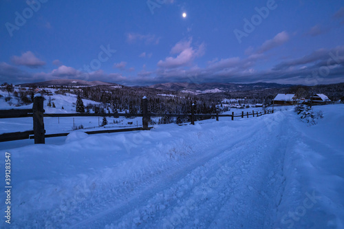 Night countryside road, hoases, hills, groves and farmlands in winter remote alpine mountain village. Ukraine, Voronenko, Moon above village.