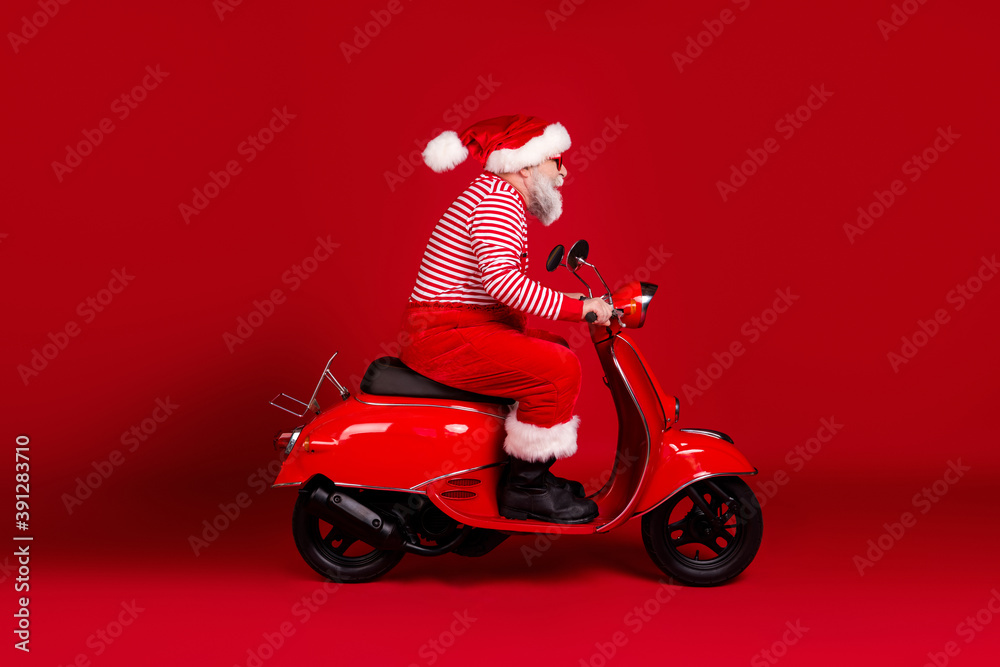 Full length body size profile side view of his he handsome bearded Santa father riding moped fast hurry delivering gift North Pole traveler isolated bright vivid shine vibrant red color background Full length body size profile side view of his he handsome bearded Santa father riding moped fast hurry delivering gift North Pole traveler isolated bright vivid shine vibrant red color background