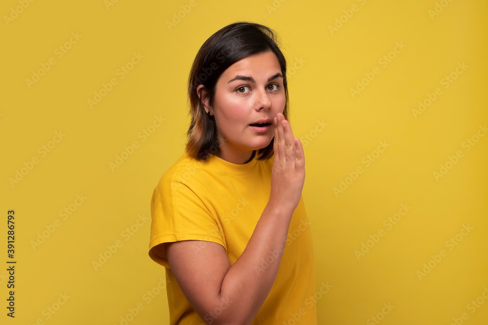Caucasian woman covering mouth with hand tells secrets to someone, isolated on yellow wall. Caucasian woman covering mouth with hand tells secrets to someone, isolated on yellow wall.