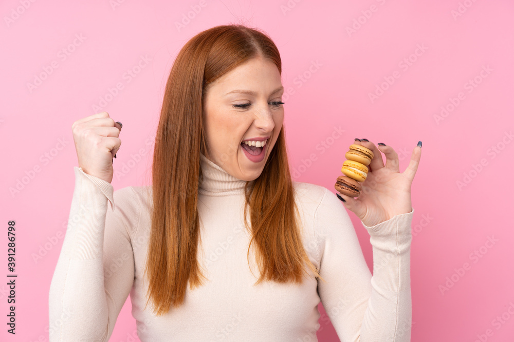 Young redhead woman over isolated pink background holding colorful French macarons and celebrating a victory Young redhead woman over isolated pink background holding colorful French macarons and celebrating a victory