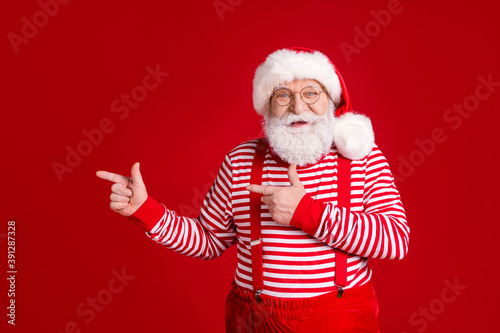Photo of retired old man white beard direct finger empty space suggest interesting solution wear santa x-mas costume suspenders spectacles striped shirt cap isolated red color background © deagreez