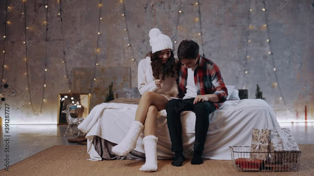 Excited young Caucasian couple enjoying winter holidays, looking at mobile phone screen shocked by seasonal sales or discount online, sitting on bed at home, a positive teenager enjoying raising her Excited young Caucasian couple enjoying winter holidays, looking at mobile phone screen shocked by seasonal sales or discount online, sitting on bed at home, a positive teenager enjoying raising her